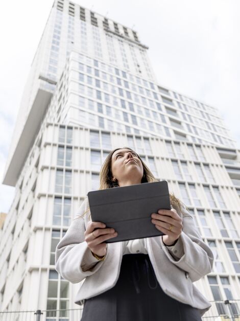Blonde woman holding iPad looking up towards white apartment building.