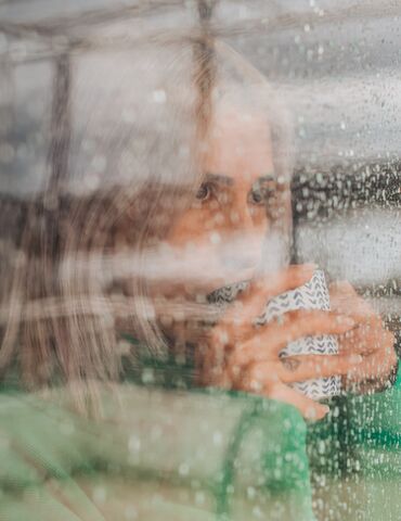 Woman drinking coffee behind glass window on a rainy day.